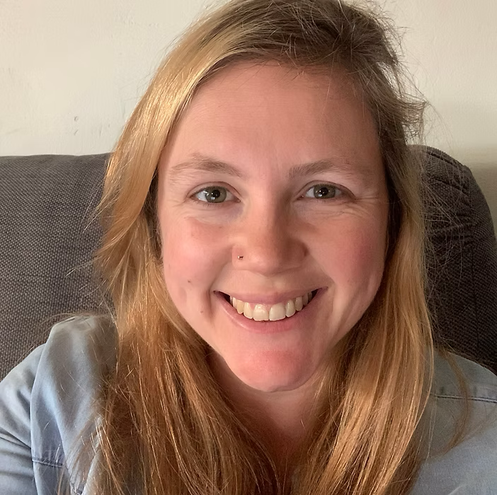 A woman with long, light brown hair smiling while sitting on a gray couch, wearing a light-colored shirt.