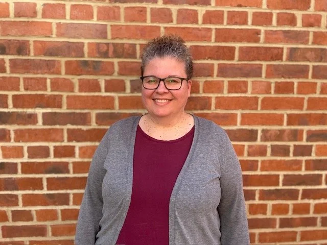 A woman with curly gray hair, glasses, and a broad smile, standing in front of a brick wall, wearing a maroon shirt and gray cardigan.