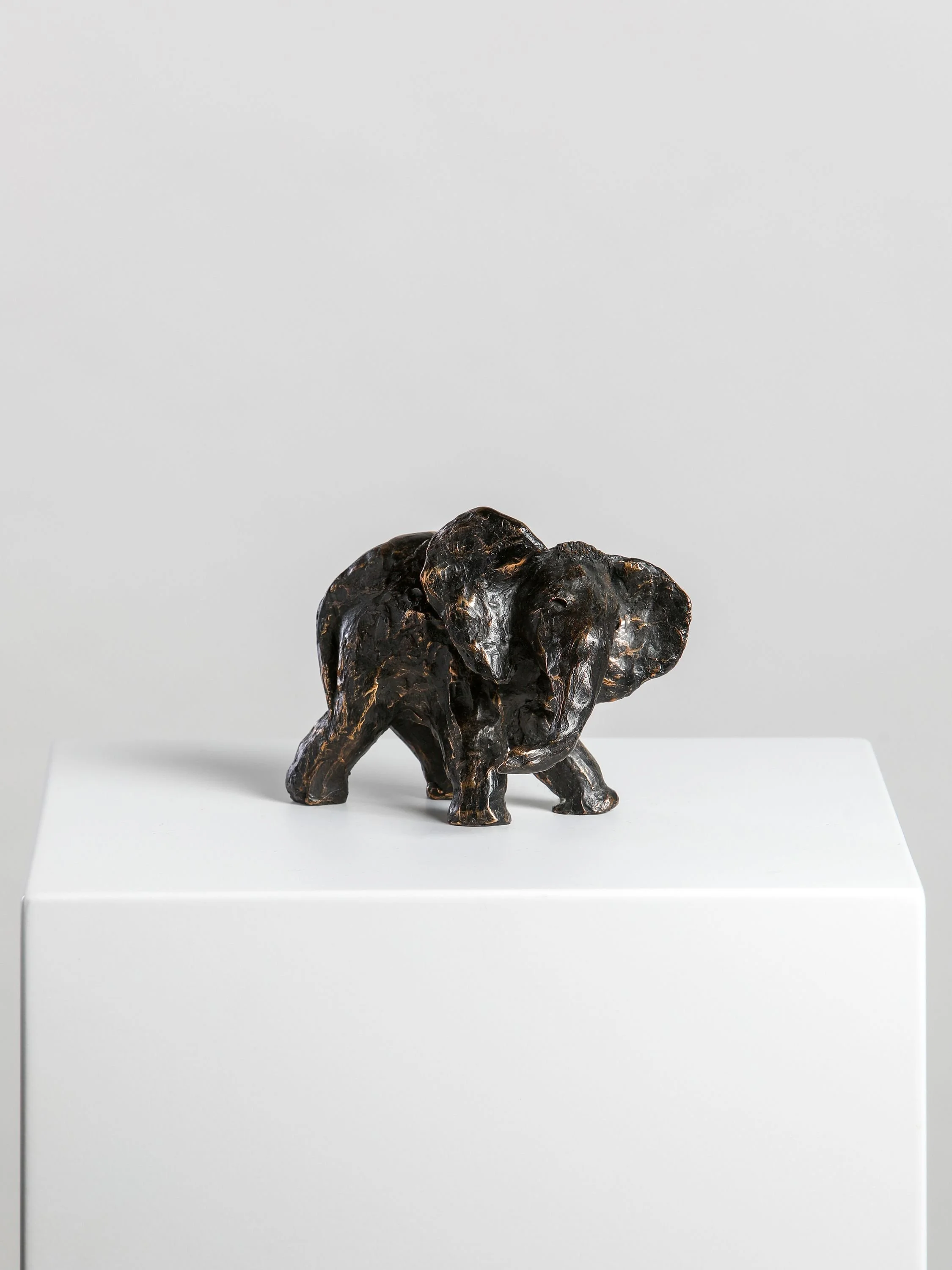 Bronze sculpture of an elephant standing on a white pedestal against a plain gray background.