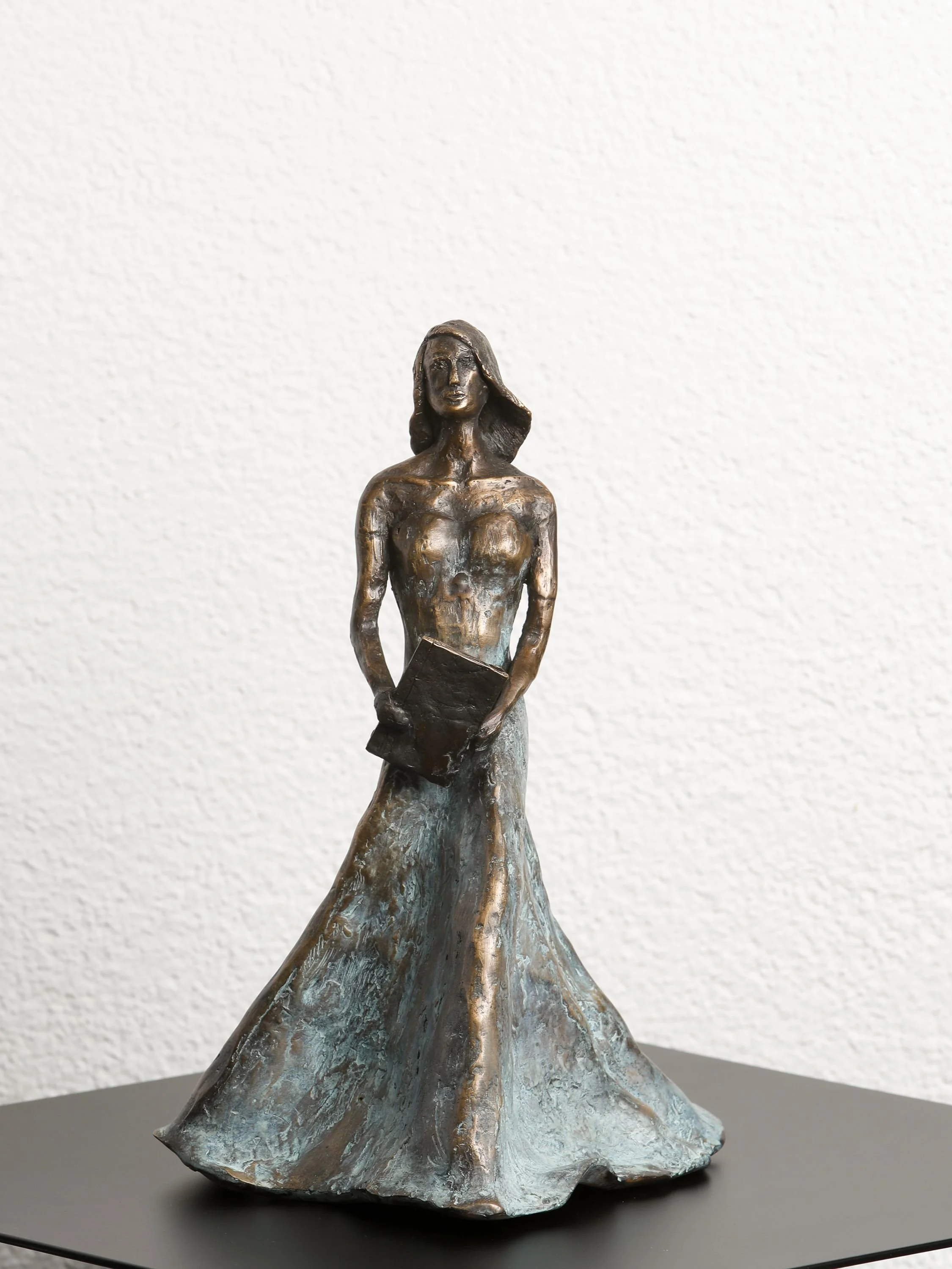 Bronze sculpture of a woman with a book, dressed in a long flowing gown, standing on a textured base against a plain white background.
