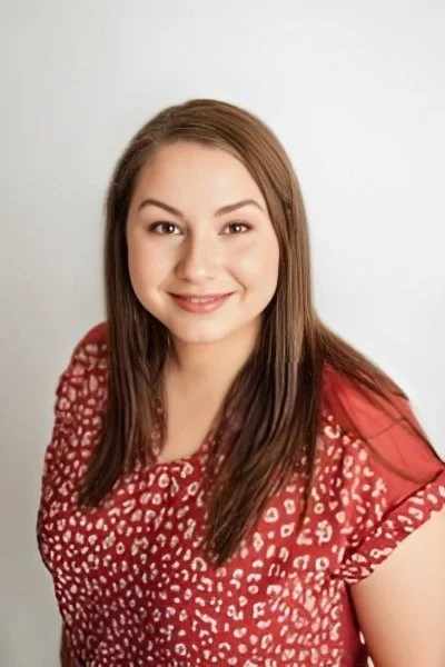 A young woman with long brown hair and a friendly smile, wearing a red patterned blouse, standing against a plain white background.
