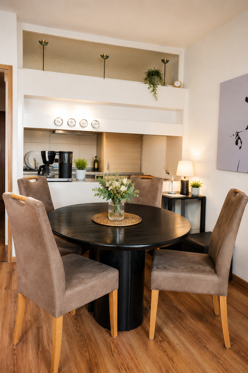 Ein Esszimmer mit einem schwarzen runden Tisch und vier beigen Stühlen. Daneben steht ein kleiner Beistelltisch mit einer Lampe und Pflanzen. Im Hintergrund ist eine offene Küche mit Kaffeemaschine, Geschirrspüler und Geschirr.