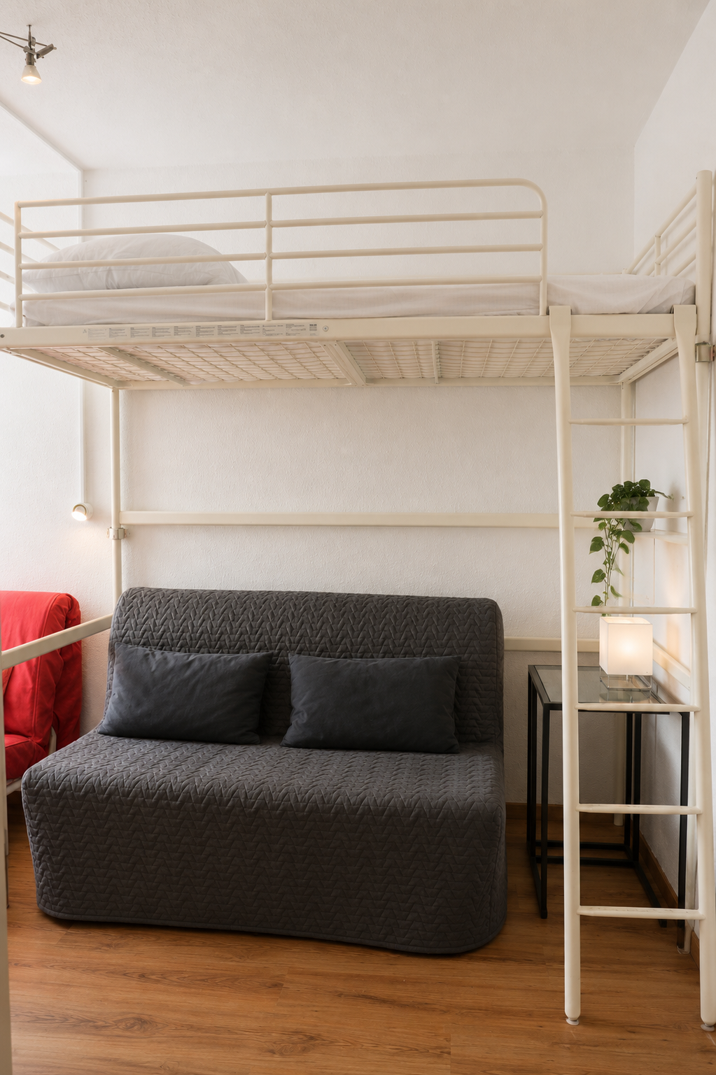 Ein Hochbett über einem grauen Sofa mit schwarzen Kissen, einem kleinen Beistelltisch mit einer Pflanze und einer Lampe, in einem gut beleuchteten Raum.