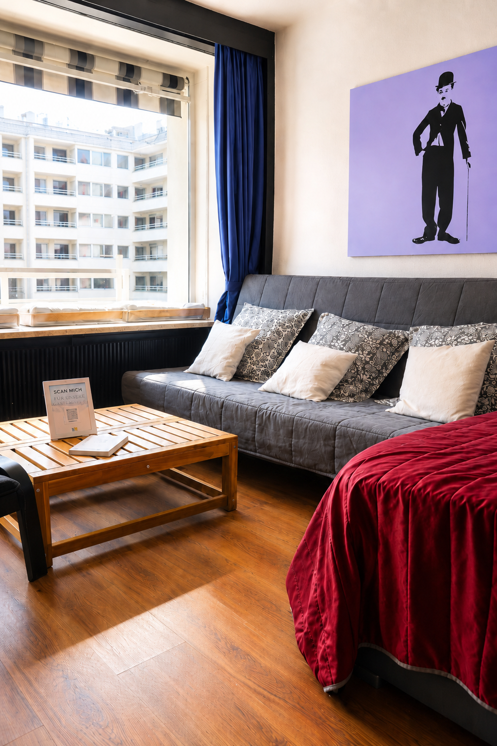 Innenansicht eines Wohnzimmers mit grauem Sofa, mehreren Kissen, einem Holztisch, einem Fenster mit blauen Vorhängen, einer modernen Kunstwand mit einem schwarzen Mann mit Zylinder, rotem Bettüberwurf und Holzboden.