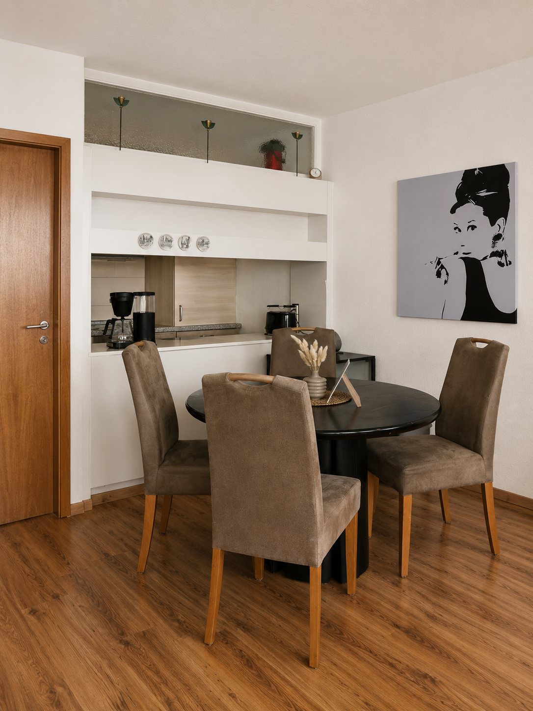 Ein Esszimmer mit einem runden schwarzen Tisch und vier braunen Stühlen. Im Hintergrund eine offene Küchenkombination, eine Wand mit Kunstwerk eines schwarzen und weißen Porträts einer Frau sowie eine kleine Dekoration auf dem Tisch.