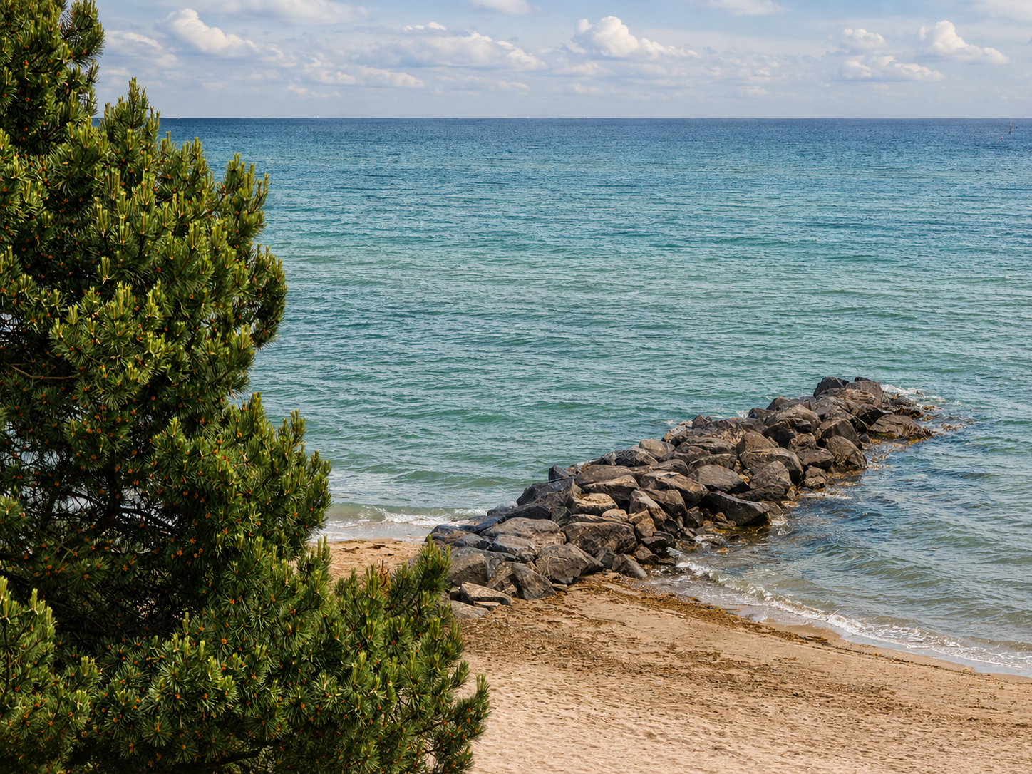 Strand mit einem Felsenpier im Wasser, neben grünen Busch, im Hintergrund Himmel mit Wolken.