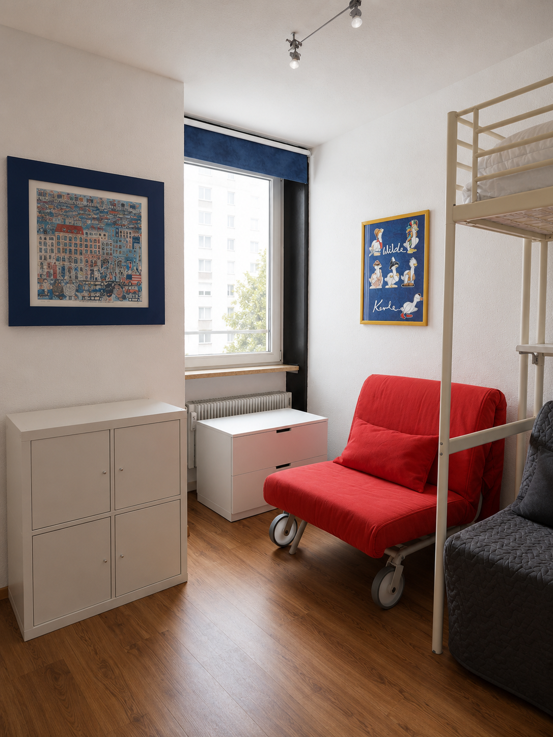 Ein kleines, hell erleuchtetes Zimmer mit weißen Wänden, Holzfußboden und einem Fenster mit blauer Jalousie. Im Raum gibt es eine rote Schlafcouch auf Rollen, einen weißen Schrank, eine kleine weiße Ablage, eine graue Couch und Hochbett. Die Wände si