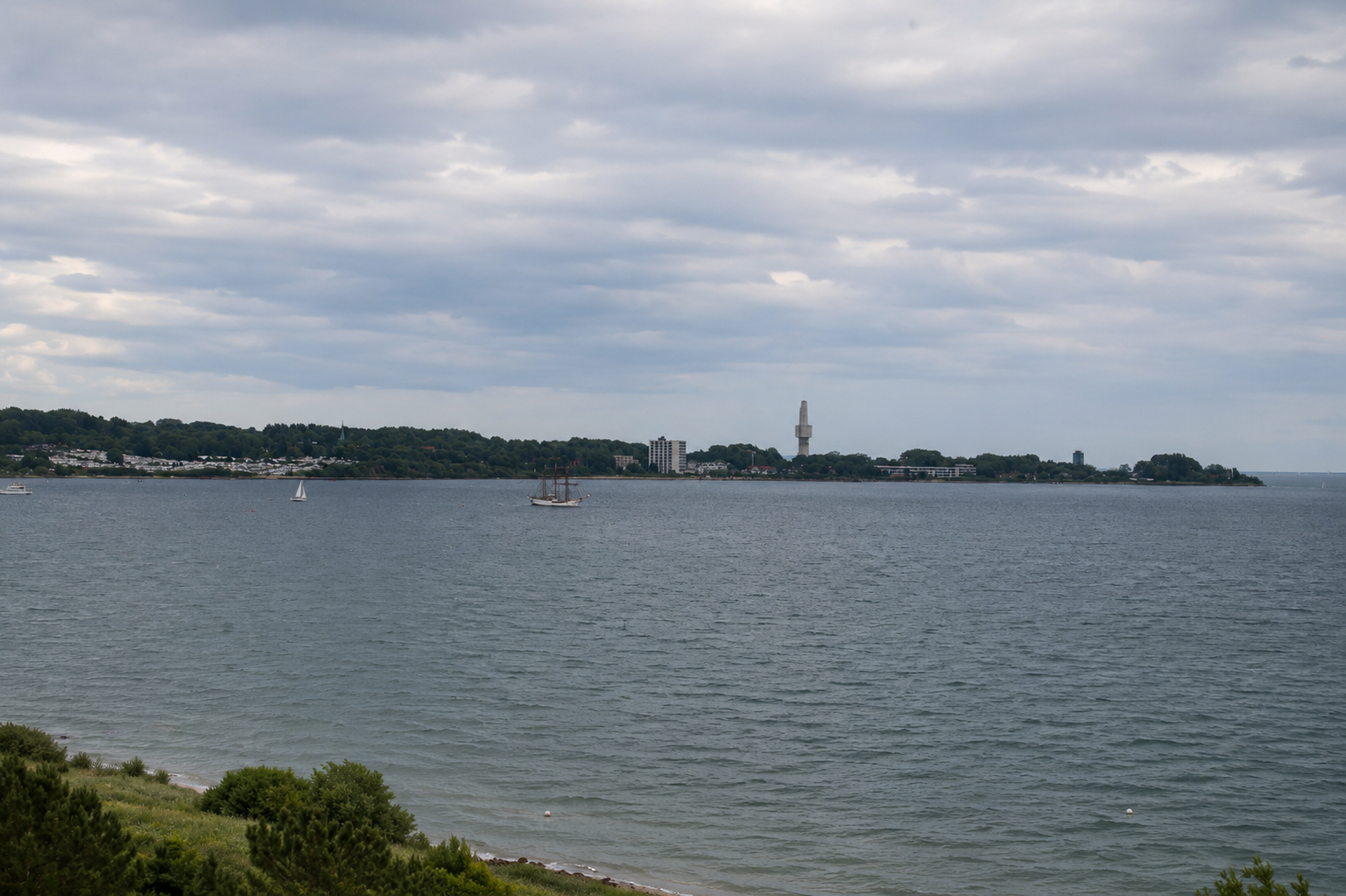 Blick auf eine Wasserfläche mit Schiffen und eine Stadt im Hintergrund, inklusive eines hohen, modernen Gebäudes, unter bewölktem Himmel.