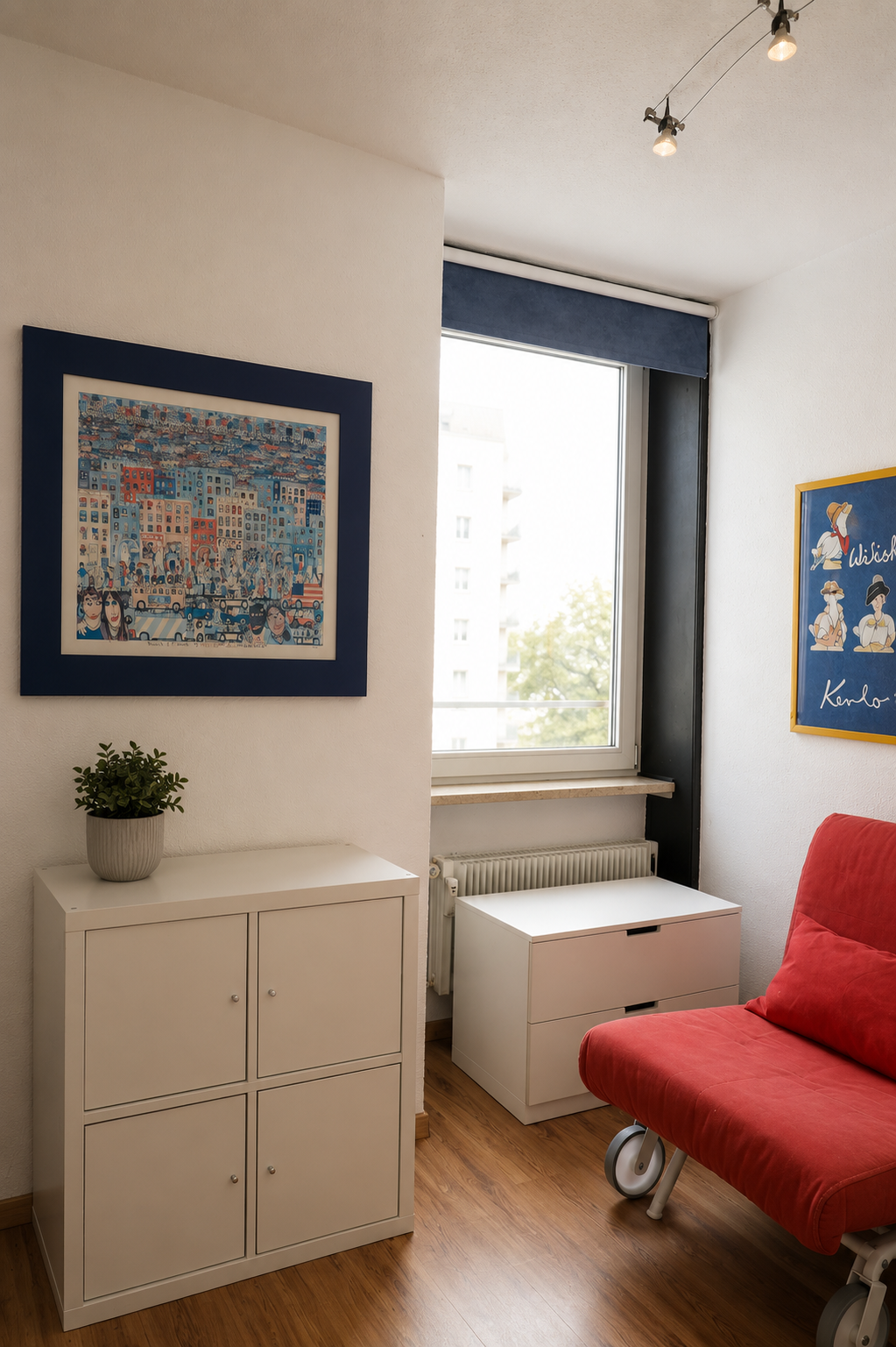 Ein helles Wohnzimmer mit weißen Wänden, einem großen Fenster, einem roten Sessel, einem weißen Beistelltisch, einem weißen Schrank mit Topfpflanze und Kunstwerken an den Wänden.