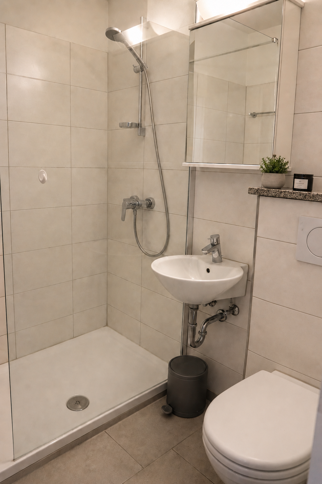 Kleines Badezimmer mit Dusche, Waschbecken und WC, beige Wandfliesen, kleine Pflanze und Kosmetikartikel auf einem grauen Regal