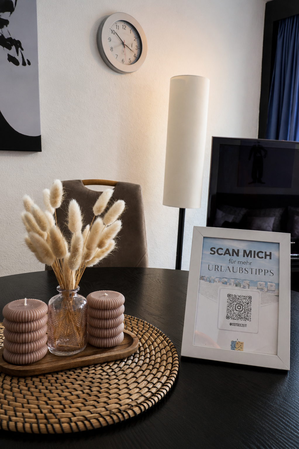 Ein dunkler Esstisch mit Kerzen, einem Blumenstrauß in einer Vase und einem QR-Code-Plakat auf einem Bilderrahmen, der für Urlaubs-Tipps wirbt. Im Hintergrund ein Stuhl, ein Stehlampe und eine weiße Wand mit einer Uhr.