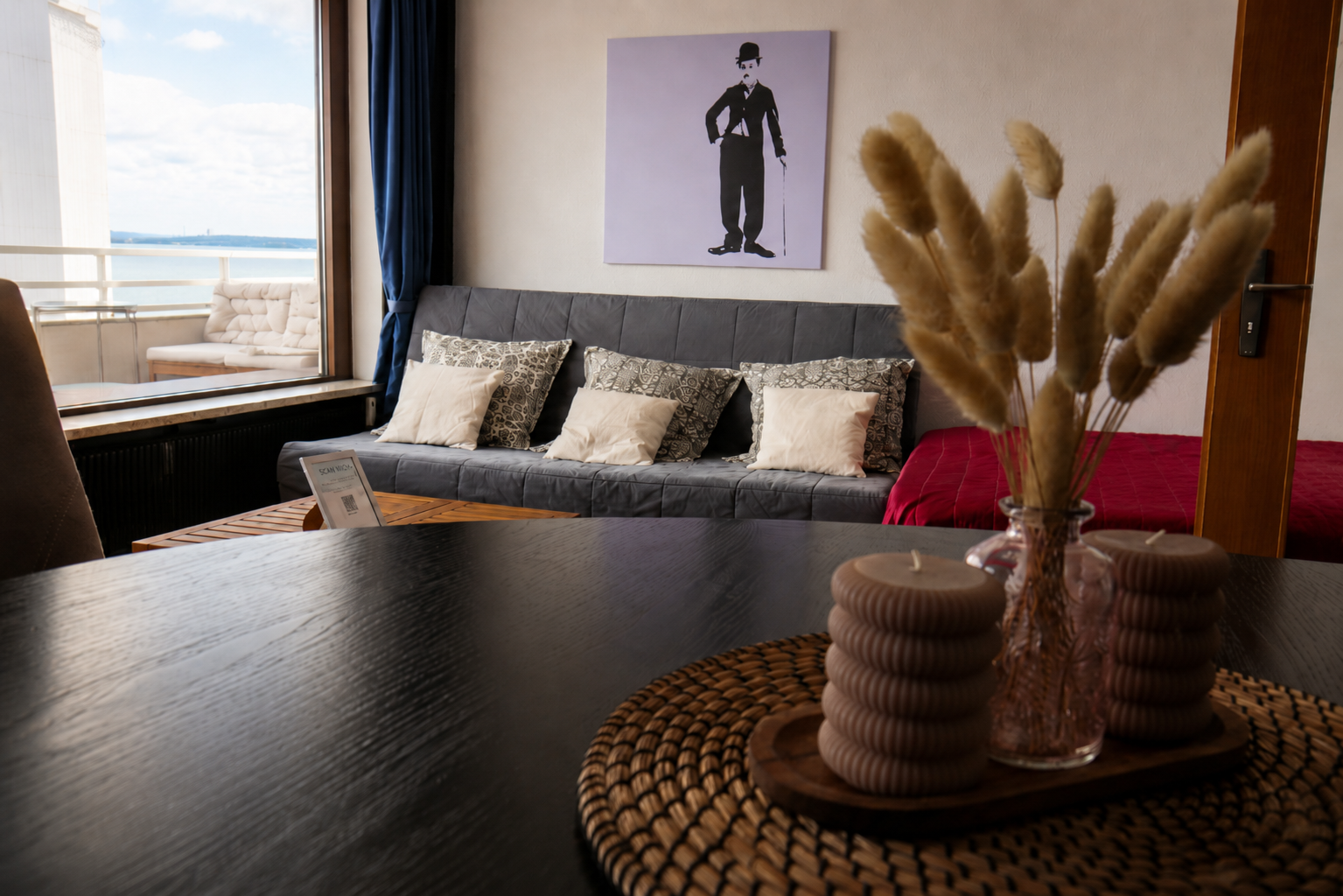 Innenraum eines Zimmers mit Couch, Kissen, Wandbild und Balkon mit Meerblick, im Vordergrund eine Vase mit Trockenblumen und Kerzen auf einem Tisch.