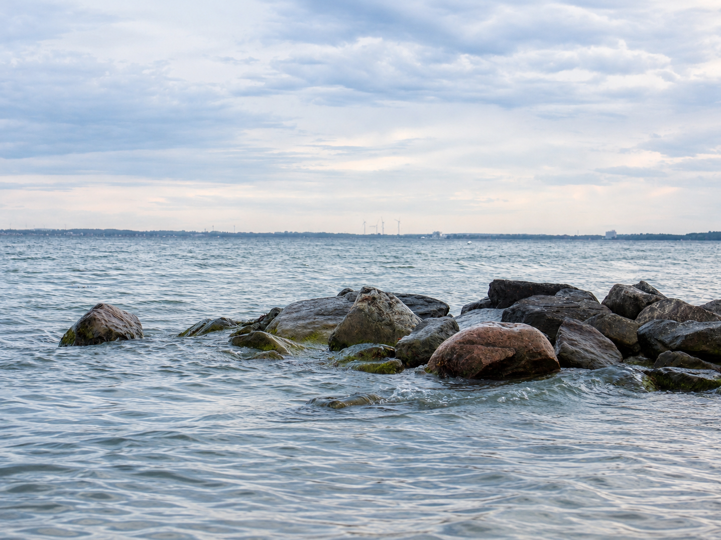 Blick auf einen See oder Fluss mit Steinen im Wasser im Vordergrund und Wolken am Himmel. Im Hintergrund sind Windkraftanlagen sichtbar.