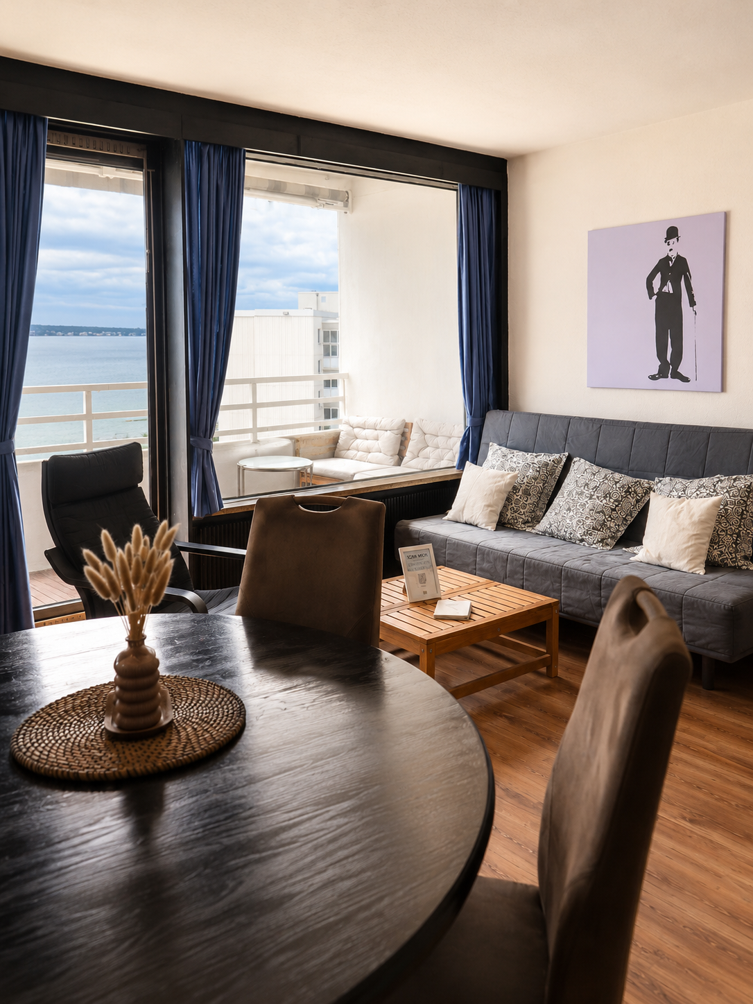 Innenraum einer Wohnung mit einem runden Holztisch, Stühlen, einem grauen Sofa mit Kissen, großen Fenstern mit blauen Vorhängen, Blick auf das Wasser und einen Balkon im Hintergrund, Wanddekor mit einer schwarzen Figur, Dekokelch auf dem Tisch.