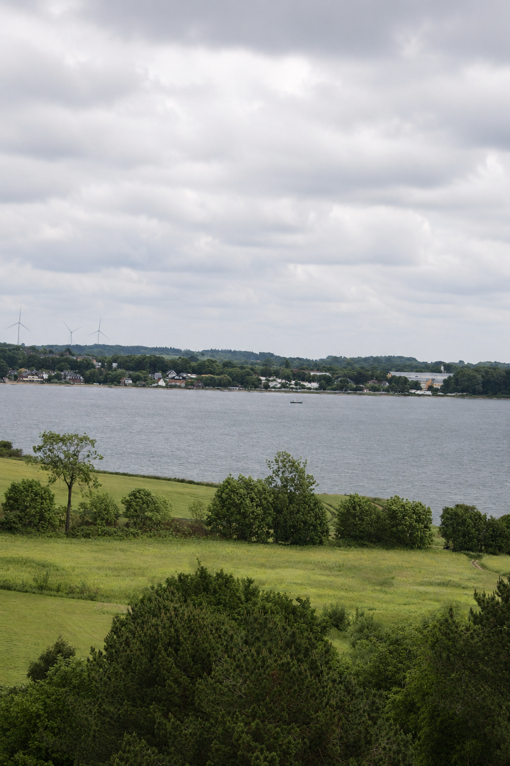Landschaft mit Wasser, grünen Bäumen, Hügeln und Windkraftanlagen unter bewölktem Himmel.
