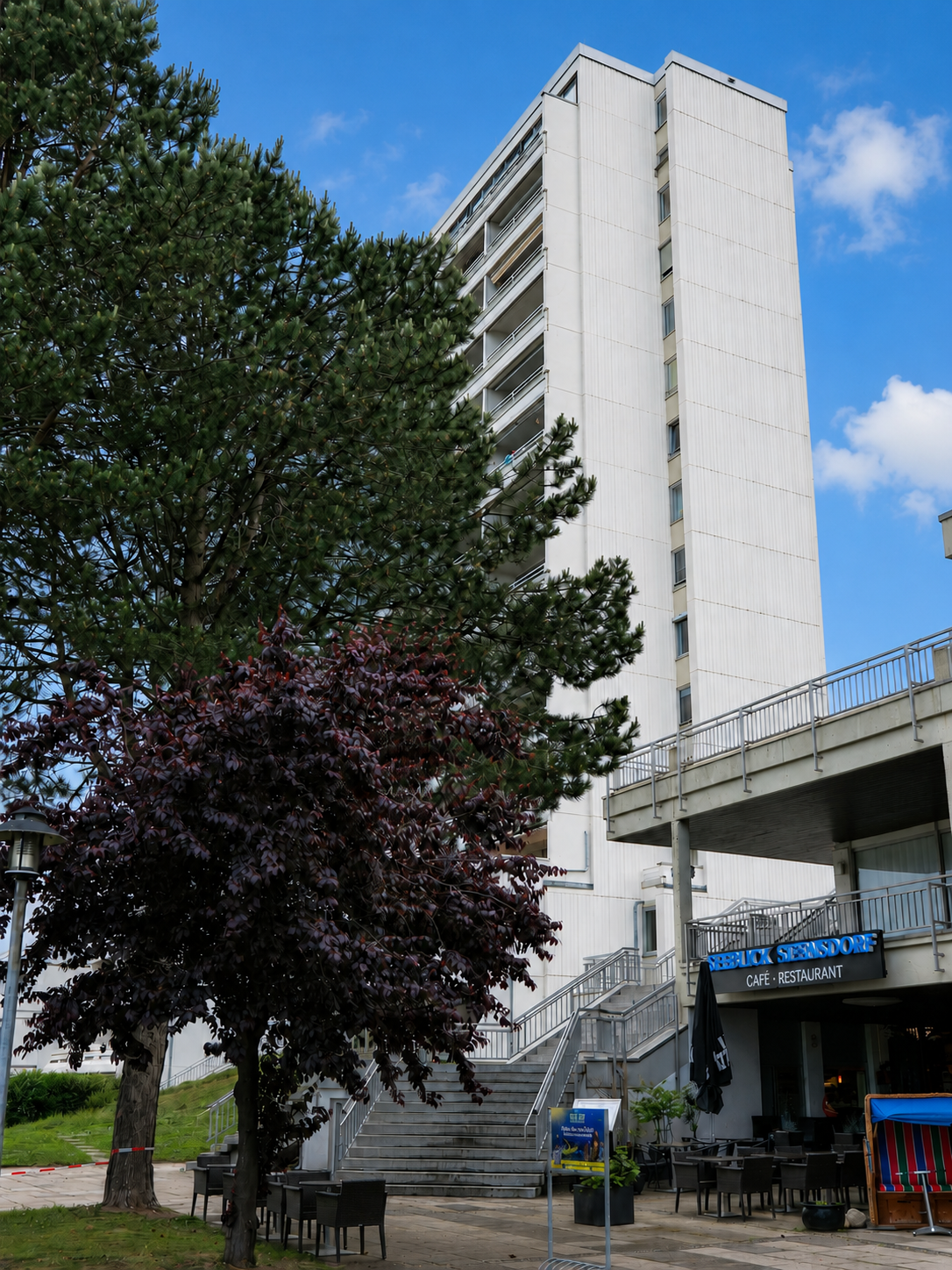 Hochhaus neben Bäumen und einem Café im Freien, blaue Himmel mit Wolken.