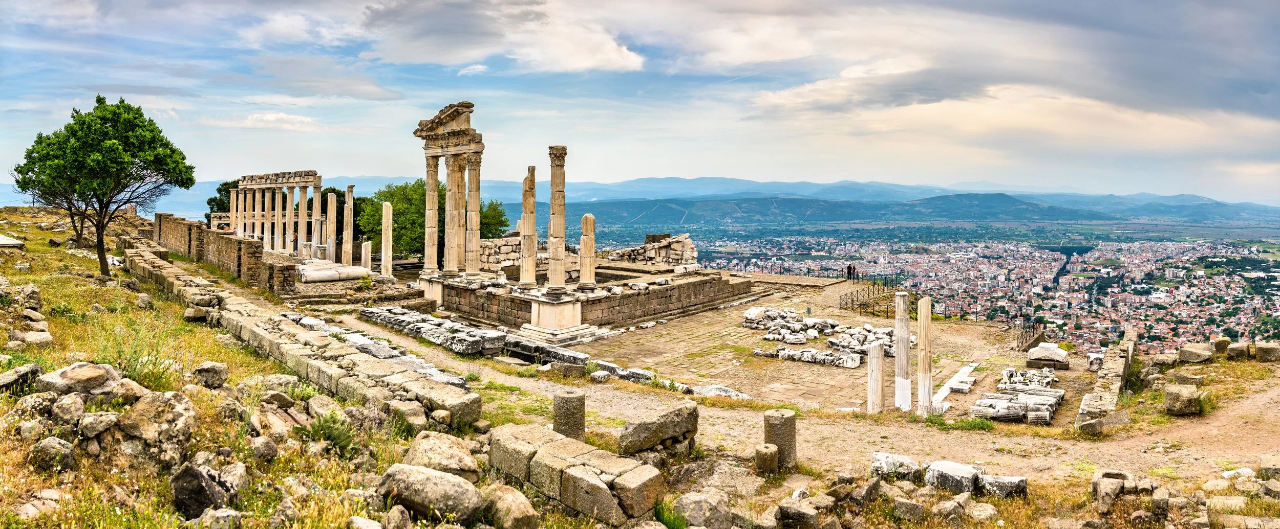 Ancient ruins with stone columns on a hilltop overlooking a city with mountains in the distance.