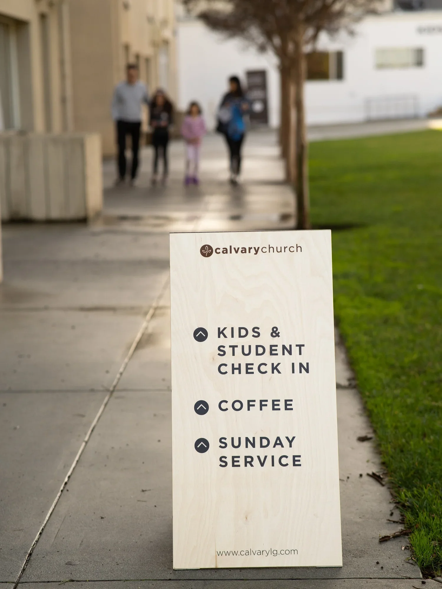 A sign outside Calvary Church with schedule details, including kids and student check-in, coffee, and Sunday service, in front of a sidewalk with a group of people walking in the background.