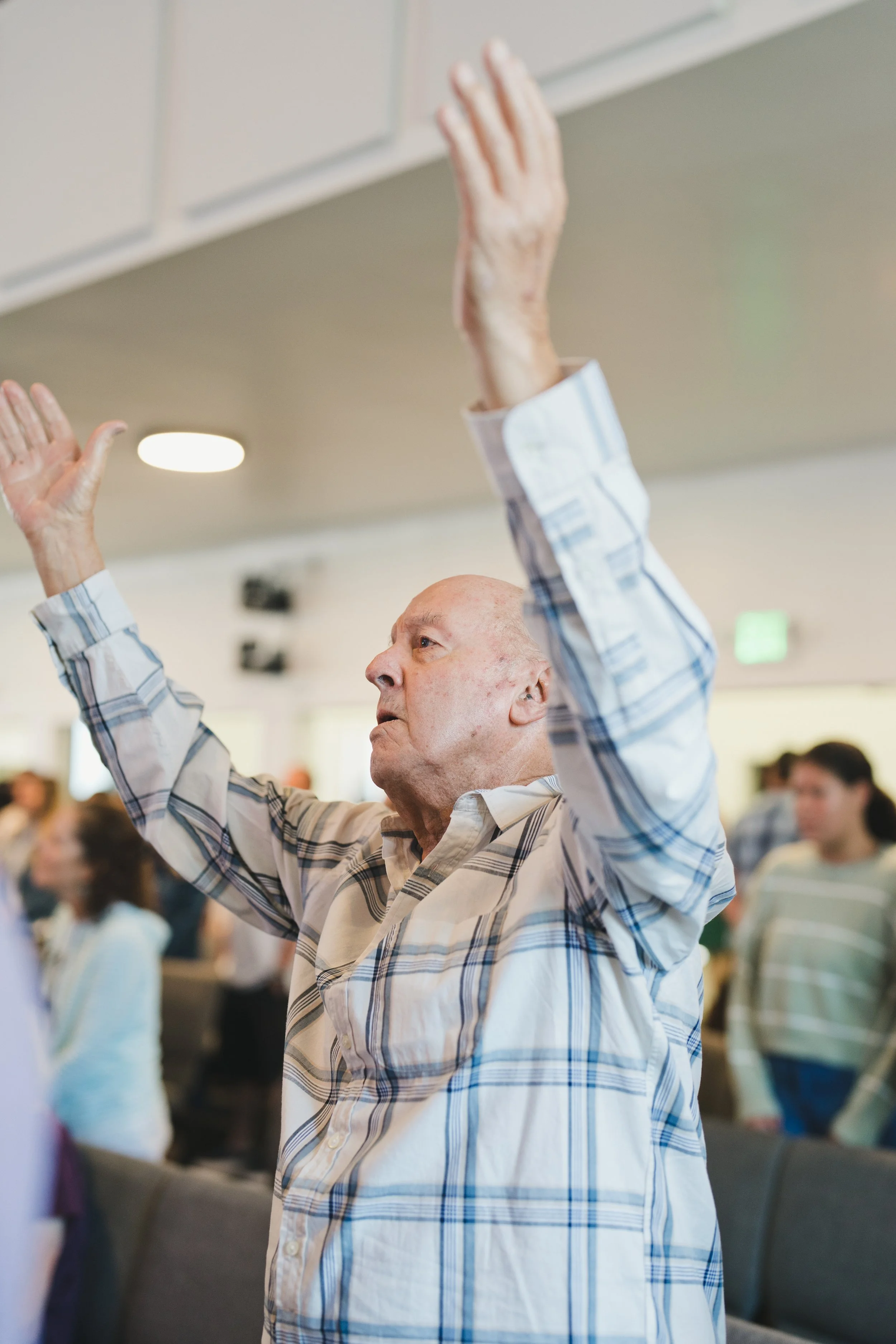 An elderly man in a plaid shirt raising his hand in a crowded indoor setting.
