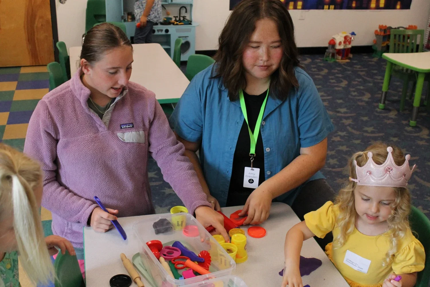 A woman with a green lanyard and name tag helping children with play dough activities in a classroom or daycare setting.