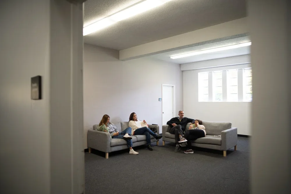 A group of five people sitting on gray sofas in a bright, minimalist room with white walls and large windows.