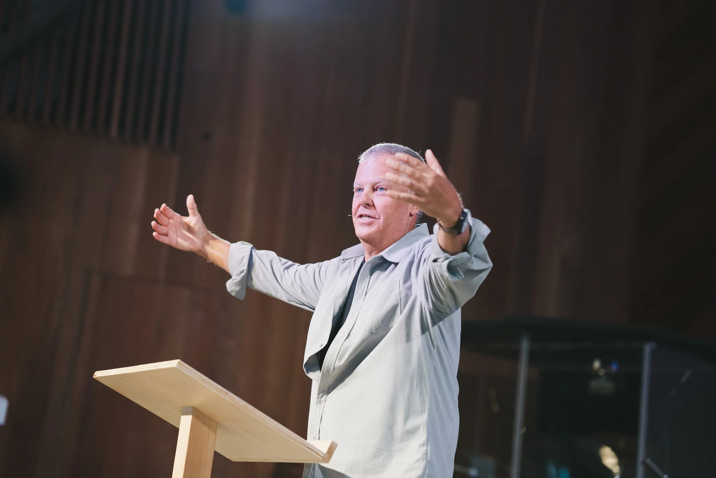 A man with short gray hair and blue eyes standing behind a wooden podium, gesturing with his arms outstretched while speaking or giving a presentation in a room with dark wood walls.