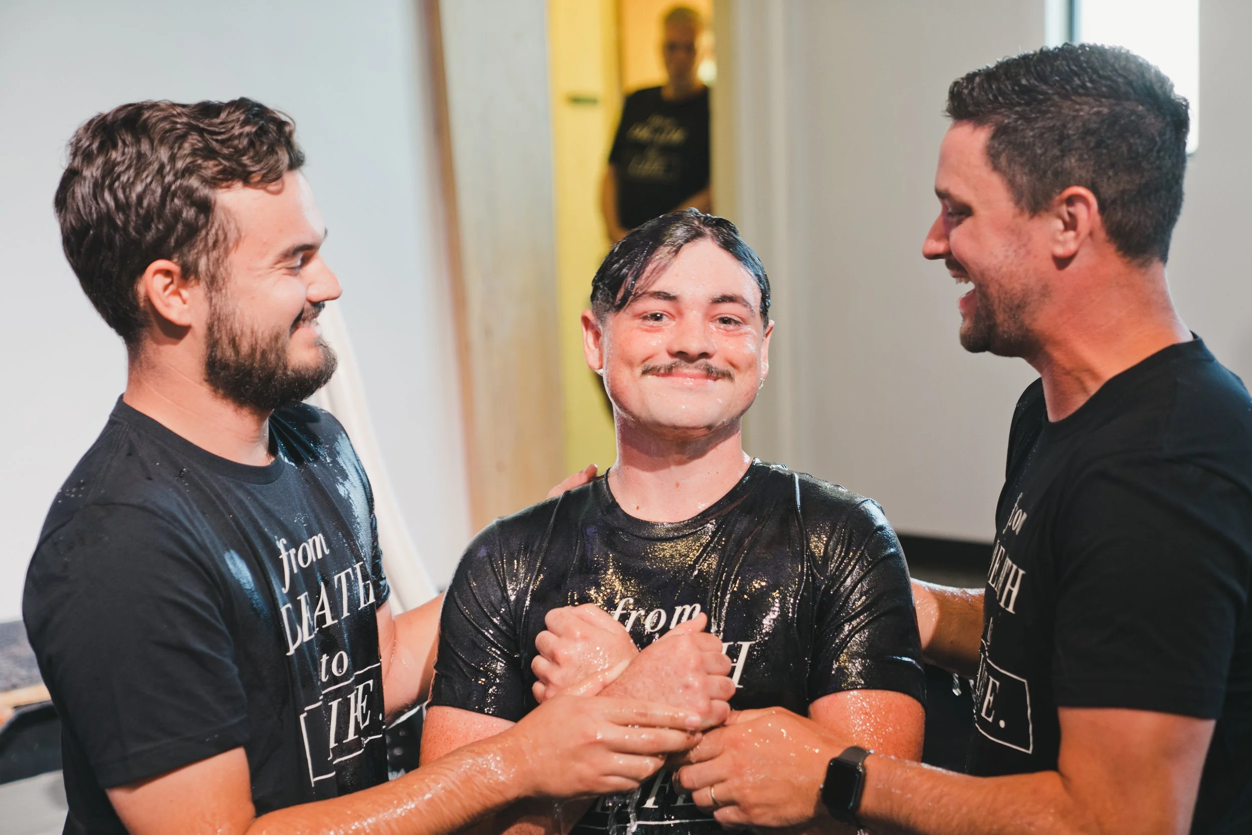 Three people smiling and holding hands in a church after a baptism or religious ceremony. The person in the middle is wet and wearing a black shirt with water and soap on their face and body, while the two people on either side are also in black shirts. One person has a beard and hair, and the other has short dark hair.