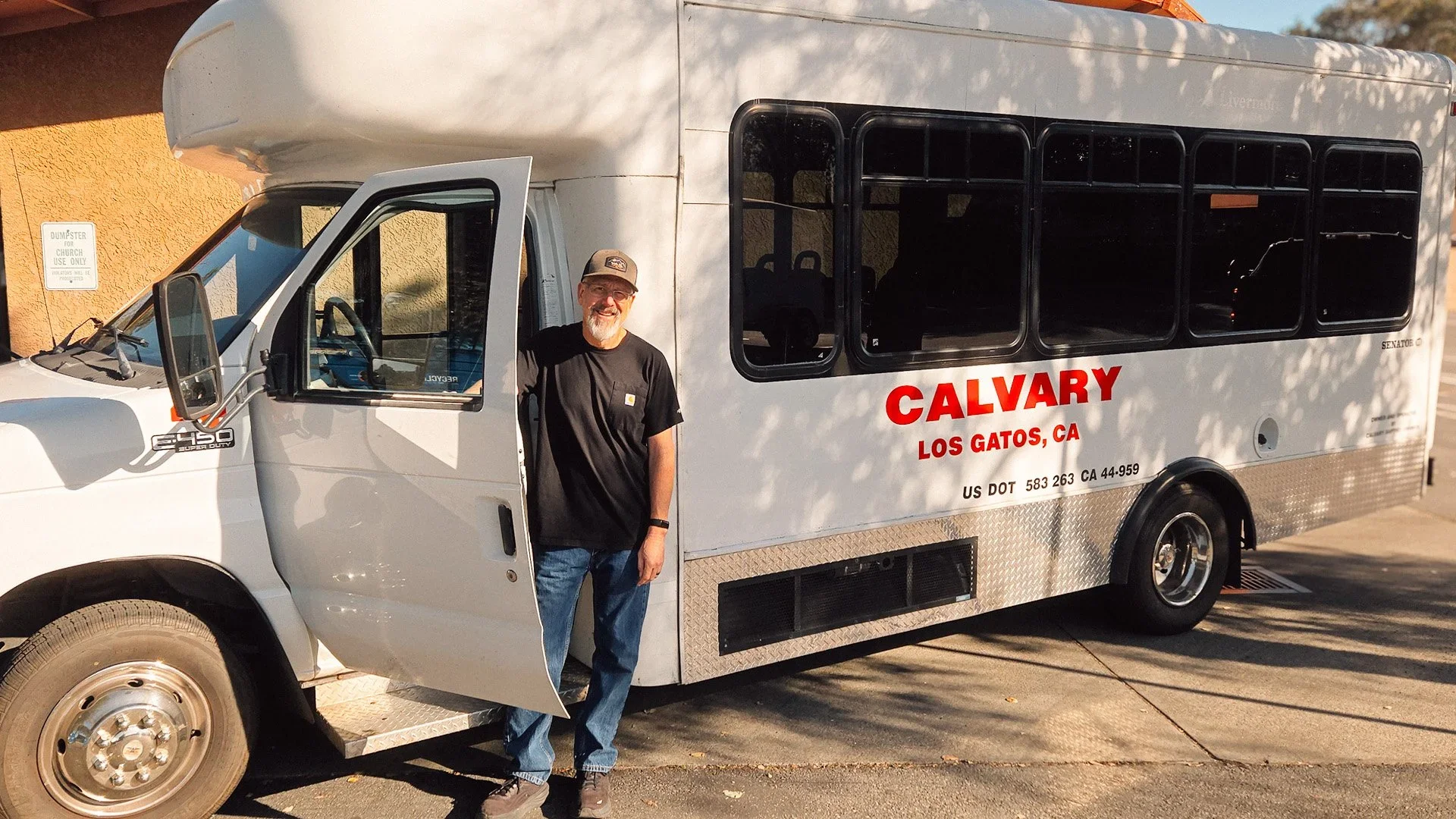 A man wearing a black T-shirt, jeans, and a baseball cap standing outside next to a white bus with large tinted windows. The bus has red text that says 'Calvary Los Gatos, CA' and includes additional vehicle information. The background includes a beige wall with a sign and shadows from nearby trees.