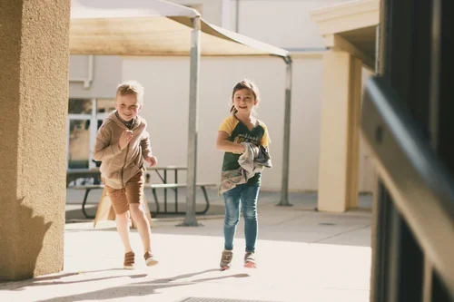 Two children running outdoors, one holding a gray jacket, under a shaded structure.