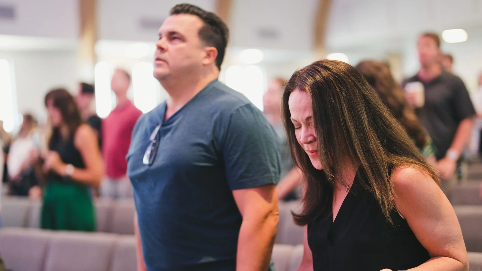 A group of people standing with eyes closed, emotionally engaged in prayer or reflection at a church service, with a man and woman in the foreground.