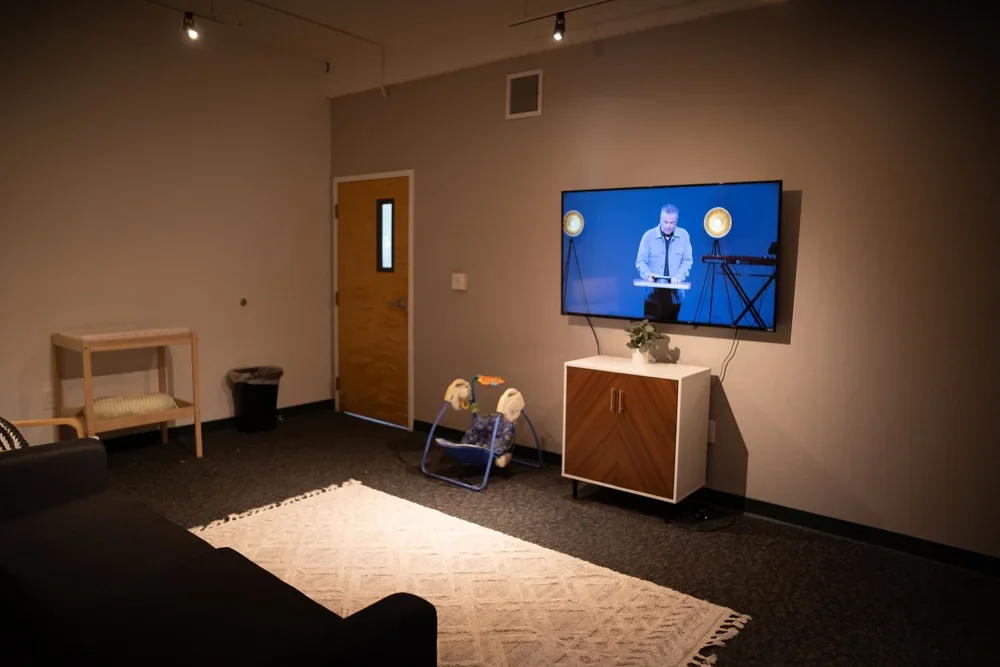 A living room with a black sofa, a white rug, a TV mounted on the wall, a small wooden table, a toy walker, and a door in the background.