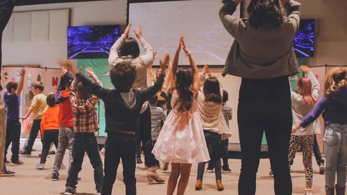 Children and adults participating in a group dance or exercise activity in a room, facing a large screen.