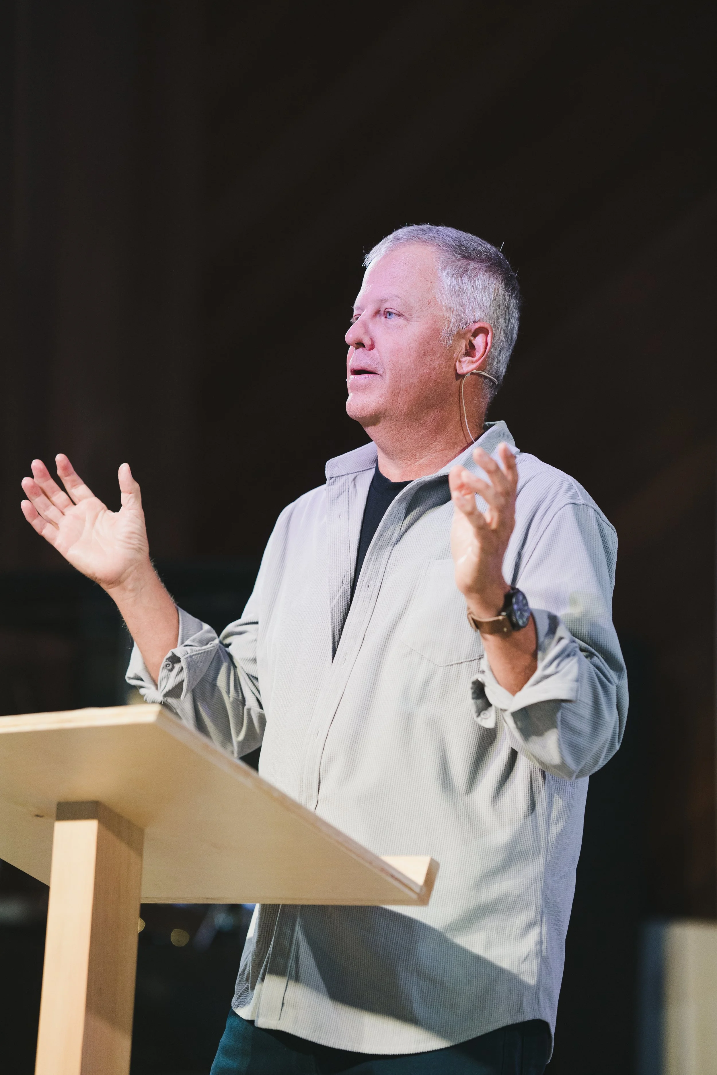 A man with gray hair wearing a headset microphone, gesturing with hands, standing at a wooden podium during a presentation or speech.