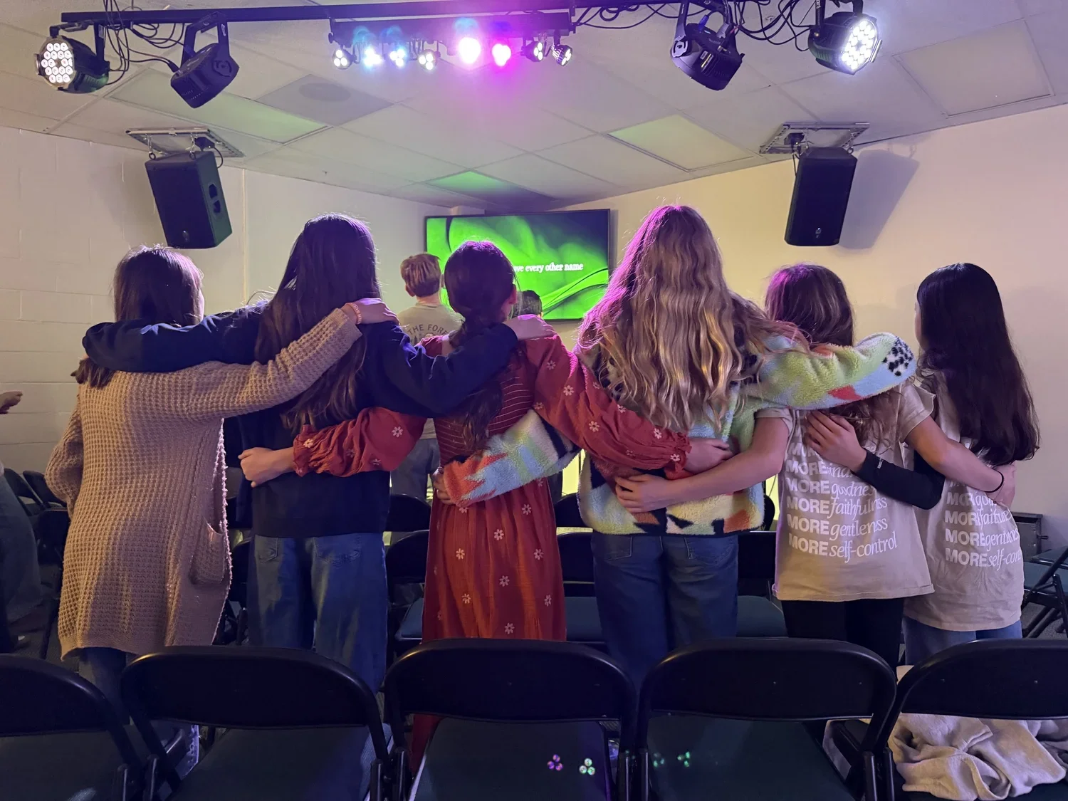 Group of young women standing in a line with arms around each other in a room with stage lighting, facing a large screen displaying green visuals.