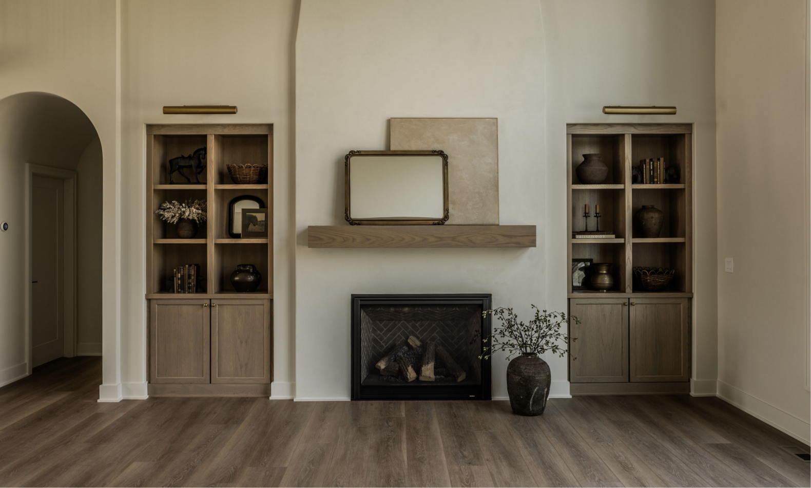 Living room with built-in wooden bookshelves on either side of a fireplace, decorative objects on shelves, a small mirror above the fireplace, and a large plant in a vase next to the fireplace.