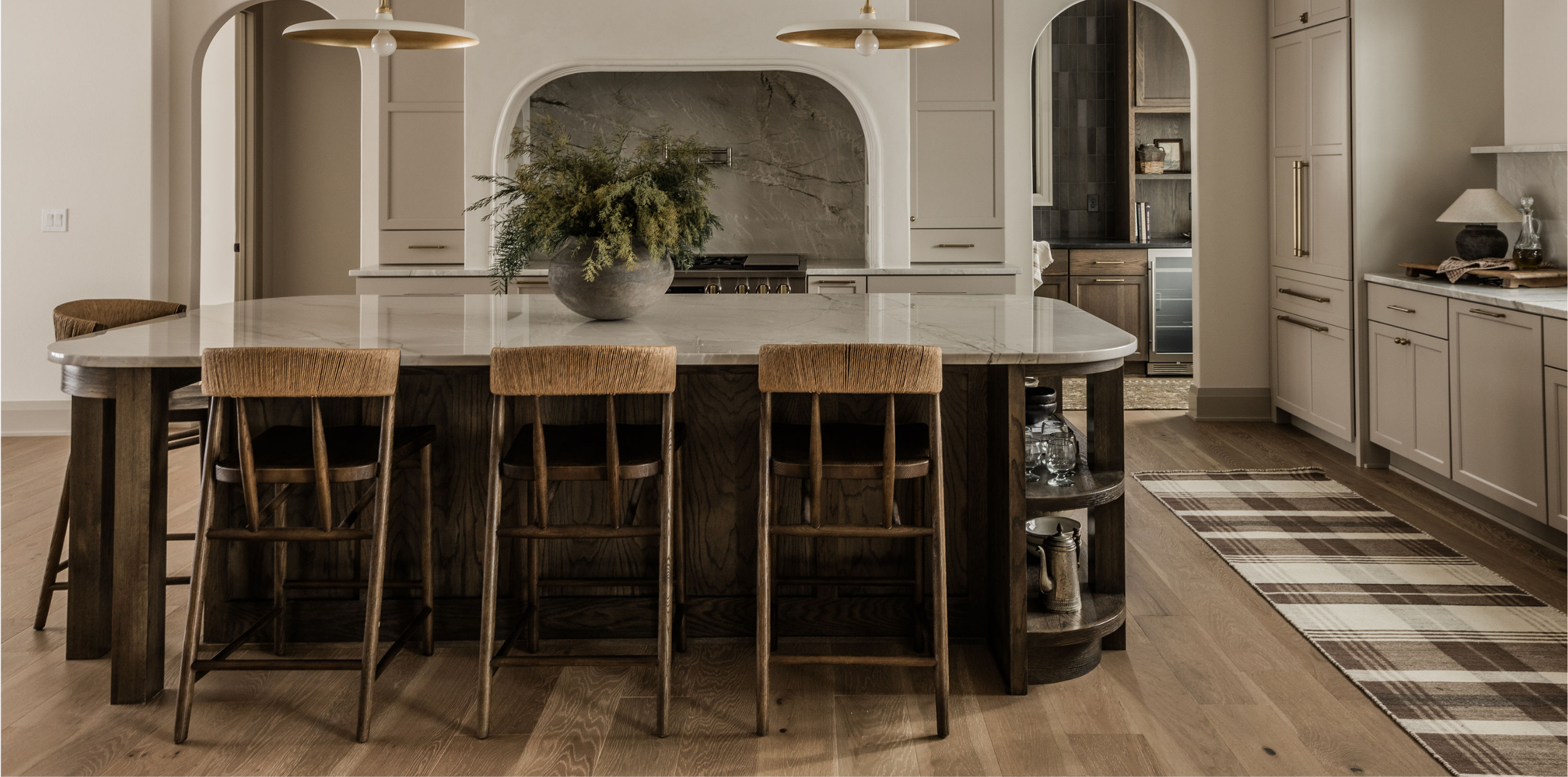 A modern kitchen with a large marble island, wooden bar stools, a large decorative vase with greenery, white cabinets, a plaid rug on hardwood floors, and a patterned backsplash.