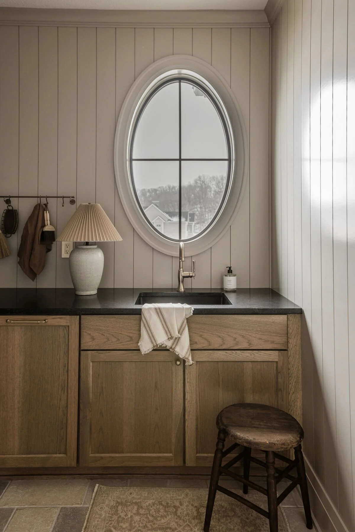 A kitchen area with a black countertop, wooden cabinets, and a white oval window showing a winter scene outside. There is a beige table lamp, a soap dispenser, and a striped dish towel hanging from the sink faucet. A stool is positioned to the right of the cabinets.