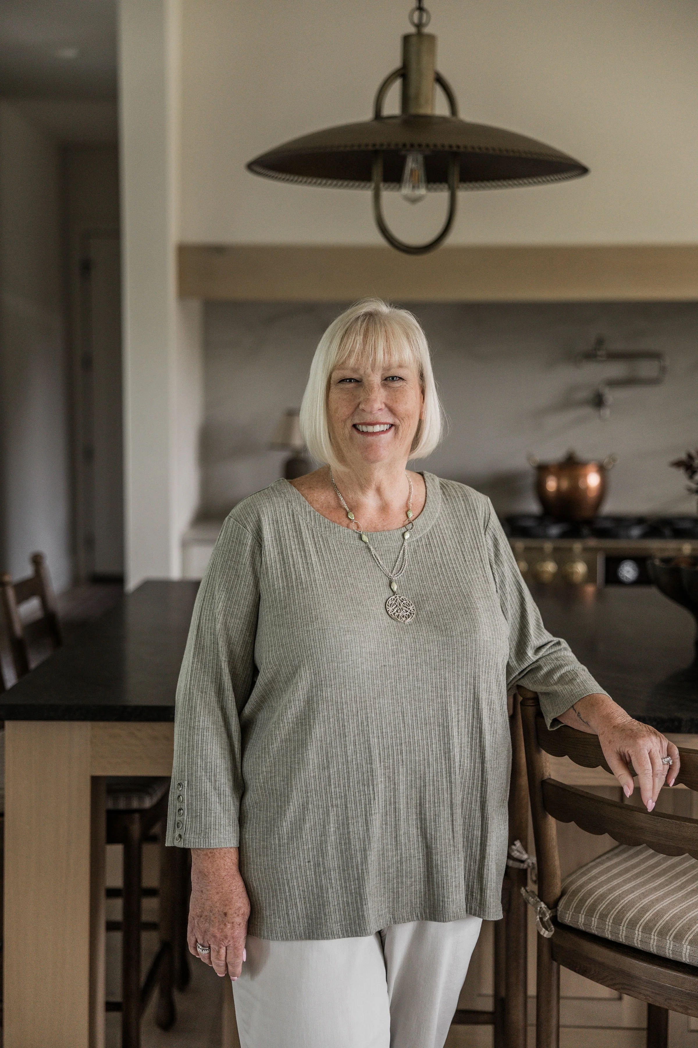 An elderly woman with short blonde hair smiling in a kitchen, wearing a beige top with a necklace, standing with her left arm resting on a wooden chair.