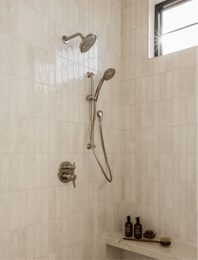 Shower with beige tiled walls, a window at the top, and bottles of shampoo or soap with brushes on a built-in shelf.