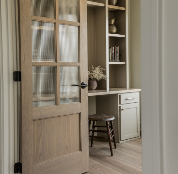 An open wooden door leads into a cozy room with a built-in desk or bookshelf, a small wooden stool, a vase with dried flowers, and books on the shelves.