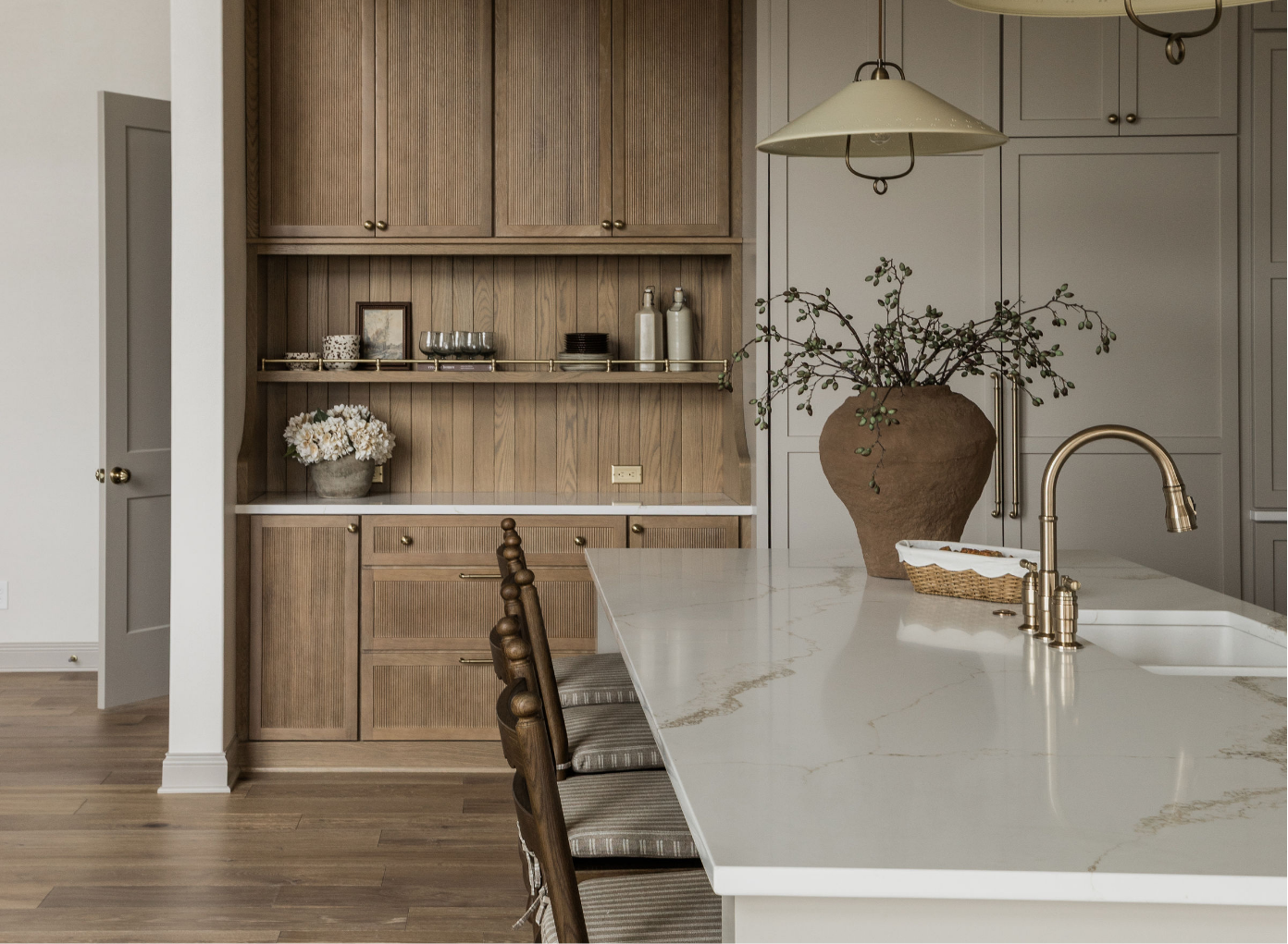 A modern kitchen with a white marble island, brass faucet, large clay vase with greenery, and built-in wooden cabinets and shelves with decorative items.