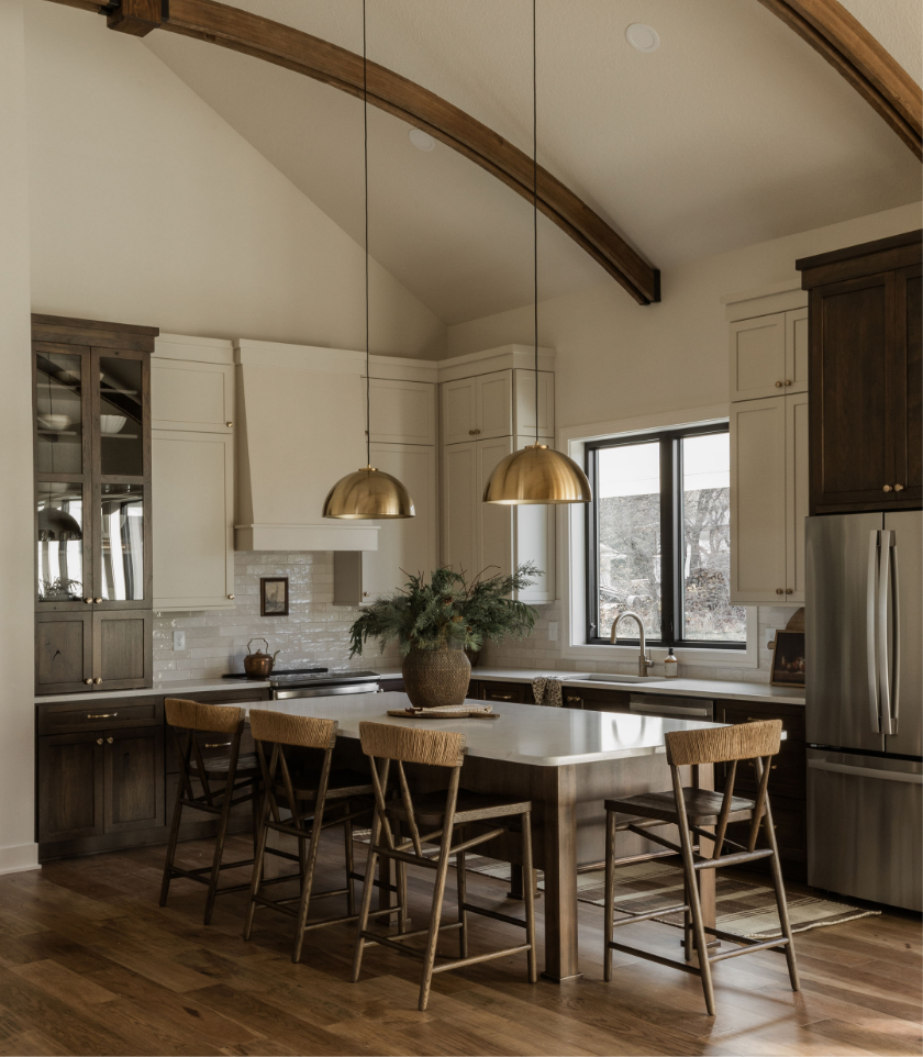 Modern kitchen with a large center island, wooden chairs, white and dark brown cabinetry, a window above the sink, and gold pendant lights hanging from a vaulted ceiling with exposed wooden beams.