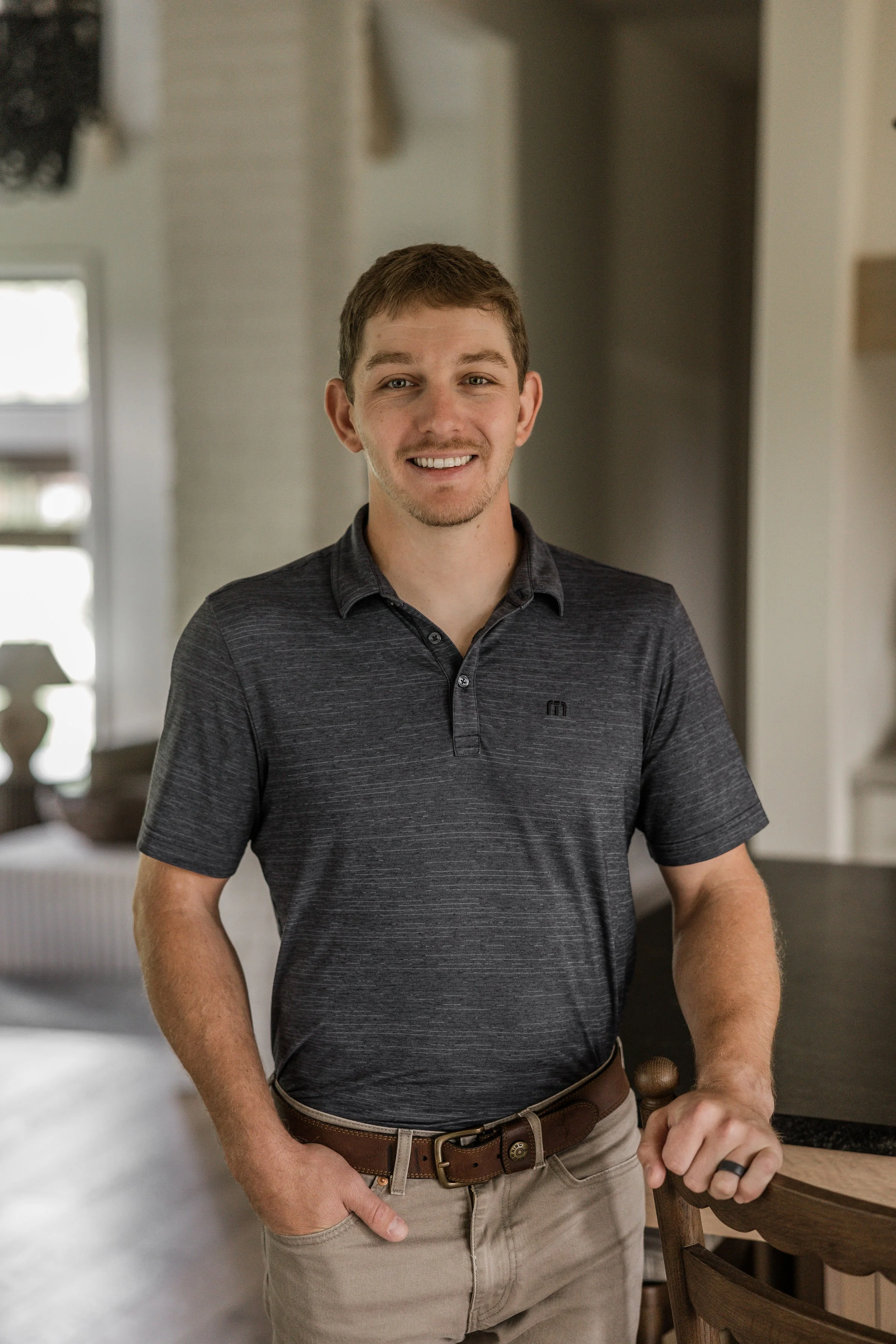 A young man with short brown hair and a light beard, smiling, wearing a dark gray polo shirt, beige pants, and a brown belt, standing in a modern home interior with blurred background, including a window and a lamp.