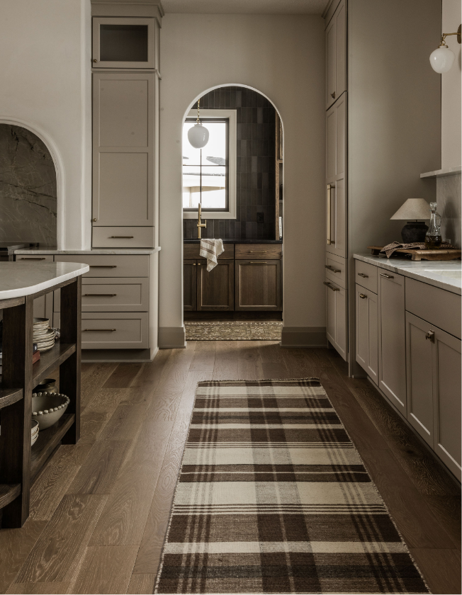 Modern kitchen with white cabinetry, wooden flooring, a plaid rug, and a small pantry or laundry area visible through an arched doorway with black backsplash.