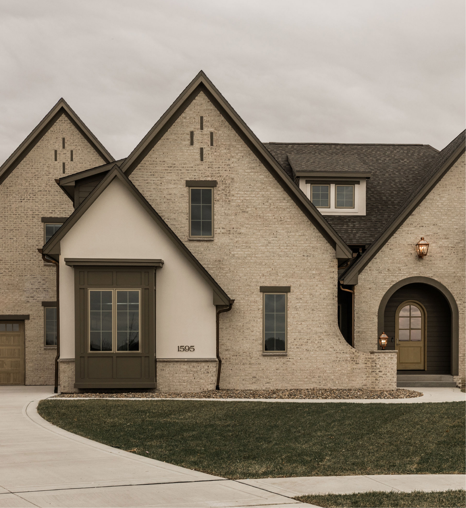 Front view of a modern house with multiple gabled roofs, brick walls, large windows, a front door with lantern lights, a well-kept lawn, and concrete pathway.