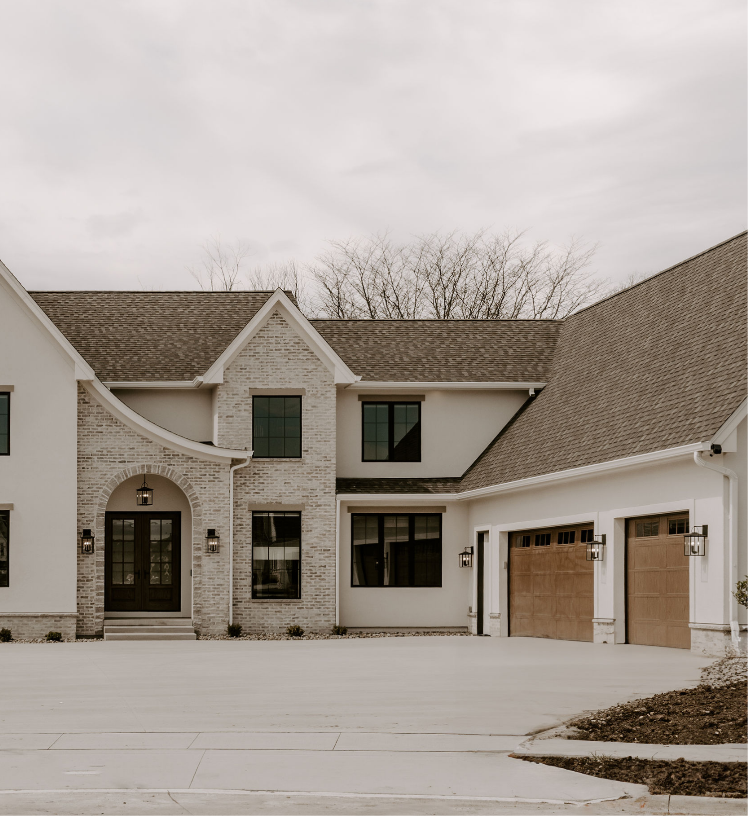 Front view of a modern house with a brick and white exterior, multiple gabled roofs with brown shingles, large windows, and a wooden garage door, with a concrete driveway and a small landscaped area.