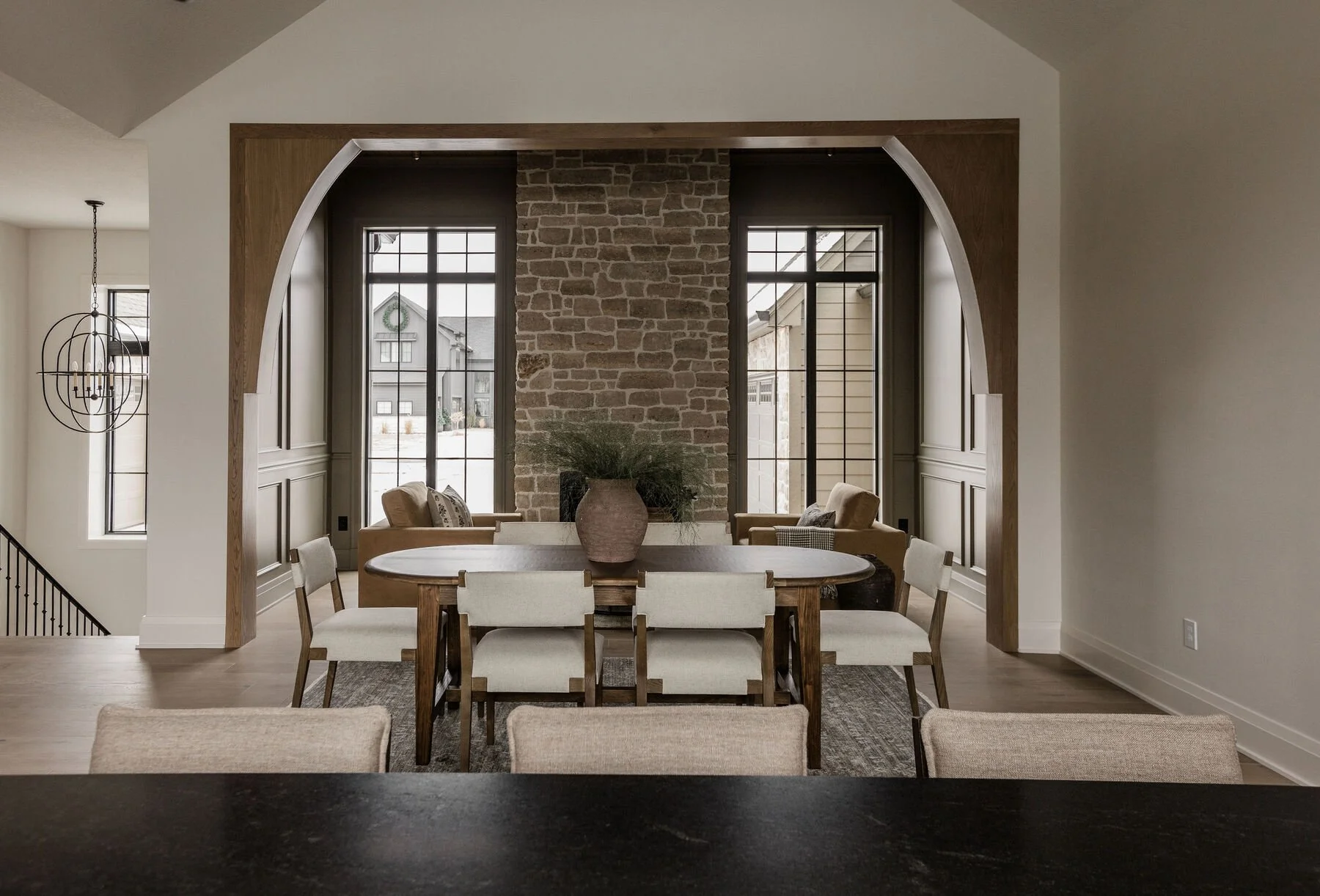 Dining room with a large oval table and eight cream-colored chairs, a vase with greenery on the table, a brick fireplace, and large windows showing outdoor houses, with a living area featuring armchairs and a sofa.
