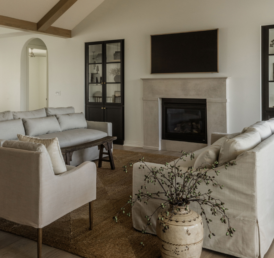 Living room with beige sofas arranged around a fireplace and a large wall-mounted TV above it, a black display cabinet with glass doors to the left, and a decorative vase with branches in the foreground.