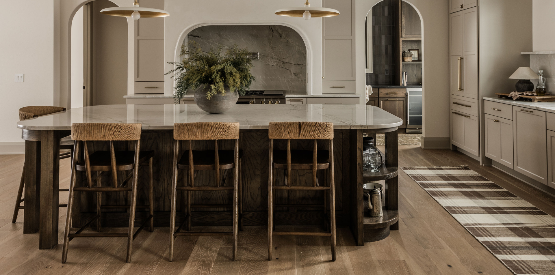 Modern kitchen with a large central island and wooden chairs, white cabinetry, a marble backsplash, and a floral centerpiece on the island. hardwood floors and a plaid rug are also visible.