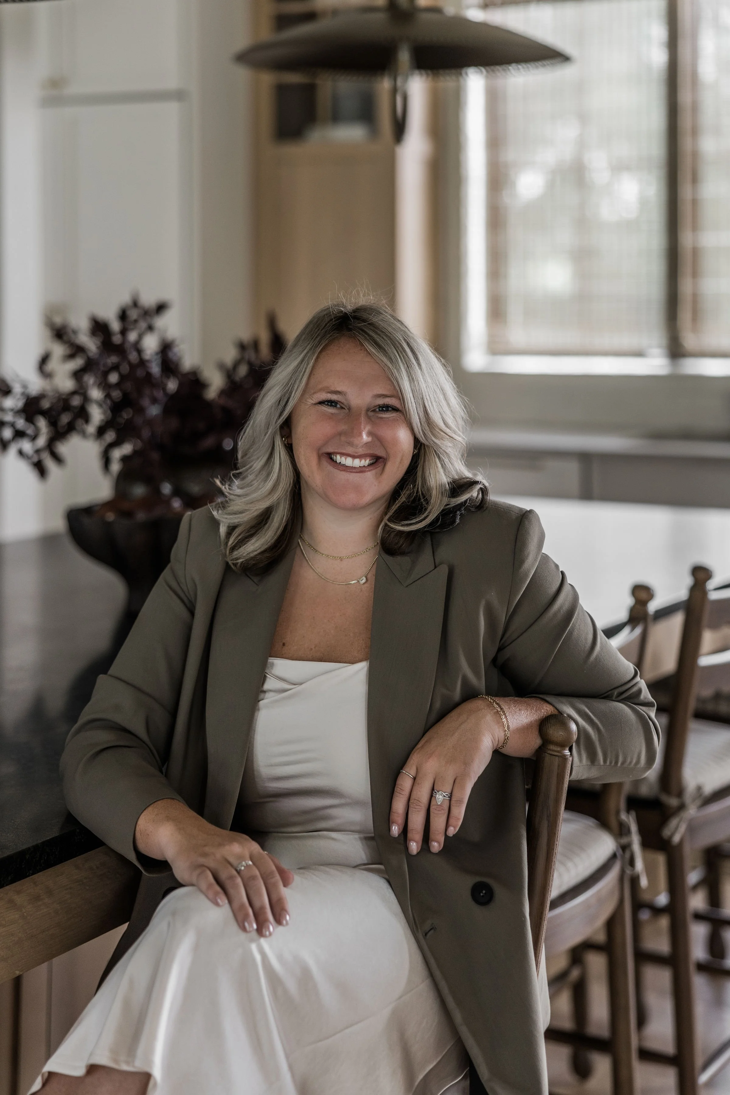 A woman with shoulder-length blonde hair, smiling and sitting at a dining table in a well-lit room with large window blinds. She is wearing a beige blazer over a white top, with jewelry including rings, necklaces, and a bracelet.